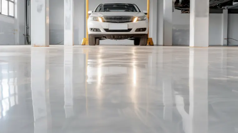 A car workshop features a glossy epoxy floor with a lifted sedan prominently displayed. Bright lighting enhances the clean and modern look of the workspace